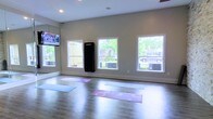 This single-floor fitness and yoga studio is thoughtfully designed to support a range of wellness and exercise activities. The open floor plan provides ample space for group or individual practice, with hardwood-style flooring that offers both durability and comfort underfoot. Several yoga mats are laid out across the floor, illustrating the room's capacity to accommodate multiple participants simultaneously.

Natural light flows generously into the space through multiple double-hung windows along the exterior wall, offering views of surrounding greenery and creating a bright, welcoming atmosphere. The neutral, light gray walls contribute to a clean and calming aesthetic throughout the room.

A large wall mirror runs along one side of the studio, a practical feature that allows users to monitor their form during workouts. A wall-mounted flat-screen TV is positioned for easy viewing, making the space suitable for guided workout sessions or fitness streaming. An upright fitness unit is also mounted to the wall, adding versatility to the studio's offerings.

Recessed ceiling lighting supplements the natural light, ensuring the space remains well-illuminated at any time of day. Overall, the studio presents a functional, modern environment well-suited for yoga, stretching, and general fitness use.