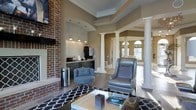 Located in Westerville, Ohio, this single-floor space showcases a thoughtfully designed interior that balances classic architectural elements with contemporary style. The open layout is anchored by a striking full-height brick fireplace, which serves as a dramatic focal point and adds warmth and texture to the main living area. Decorative white columns frame the transitions between spaces, lending a refined, formal character to the floor plan.

The tray ceiling with crown molding detail adds vertical interest and a sense of elevated sophistication throughout the main area. Warm taupe walls complement the rich hardwood-style flooring, creating a cohesive and inviting atmosphere. A bold black-and-white geometric area rug defines the seating area, which is furnished with a mix of blue and gray accent chairs arranged around a glass coffee table.

A built-in wet bar or beverage station is positioned along one wall, complete with dark cabinetry, pendant lighting, and a textured tile backsplash—ideal for entertaining. Natural light flows in through arched windows visible in the adjoining space, enhancing the overall brightness and airy feel of the interior.