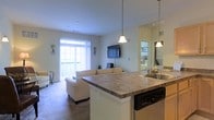 This single-floor residence is thoughtfully designed around an open-concept layout that brings together the kitchen, dining, and living areas into one cohesive and functional space. The kitchen anchors the layout with a peninsula countertop finished in a warm granite-style surface, offering casual seating and bar-style dining. A stainless steel sink, dishwasher, and pendant lighting overhead complete the kitchen's practical yet stylish setup.

The living area flows naturally from the kitchen, bathed in natural light that pours in through a large sliding glass door leading to a private balcony. Vertical blinds provide flexible light control while maintaining an airy, open feel. The neutral color palette throughout — from the soft carpeted flooring to the light-toned walls — creates a calm and welcoming ambiance.

A wall-mounted flat-screen television serves as a focal point in the living space, while a glimpse through a hallway hints at additional rooms beyond, suggesting a well-organized and private sleeping quarters. Pendant lighting and recessed ceiling fixtures ensure the space is well-lit at any hour. Overall, this residence combines comfort, functionality, and a clean modern aesthetic in a bright and inviting single-level floor plan.