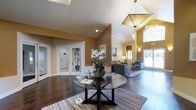 This single-story lobby makes an immediate impression with its dramatic vaulted ceilings that amplify the sense of openness and grandeur. The warm golden and taupe wall tones create an inviting atmosphere, complemented by rich, dark hardwood flooring that runs throughout the space. A striking geometric pendant light fixture serves as a focal point overhead, adding a contemporary yet refined design element.

At the center of the lobby, a circular glass-top table anchors the space, adorned with a fresh floral arrangement that enhances the welcoming ambiance. A striped area rug defines the central gathering zone, while a comfortable seating area with upholstered chairs and a sofa is positioned nearby, providing a relaxed space for visitors.

Natural light pours in through large windows and a glass door on the far end, offering views of lush outdoor greenery. Recessed lighting supplements the natural illumination throughout. White-framed French doors lead to additional areas of the building, adding architectural character. Wall sconces provide warm accent lighting, contributing to the overall polished and hospitable character of this well-appointed lobby.