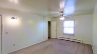 This single-story space features a well-proportioned room finished with wall-to-wall carpeting in a neutral tone, contributing to a clean and understated aesthetic. The room is illuminated by a ceiling fan with an integrated light fixture, providing both air circulation and overhead lighting. A single window allows natural light to filter into the space, brightening the otherwise simple, cream-colored walls. A baseboard heating unit runs beneath the window, offering an efficient heating solution suited for cooler climates. A closet with a sliding door and visible hanging rod provides practical built-in storage. The room's straightforward layout and neutral finishes offer a versatile canvas adaptable to a variety of uses, whether as a bedroom or a flexible living space. The overall ambiance is modest and functional, with minimal ornamental detail, making it an easy space to personalize.