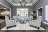 Located at 1717 Lavista Rd NE in Atlanta, Georgia, this single-floor space presents a polished and sophisticated interior designed with refined architectural details throughout. Rich hardwood flooring flows seamlessly across the open layout, complemented by neutral gray walls, crisp white crown molding, and coffered tray ceilings that lend an elevated sense of height and grandeur.

The main lounge area serves as the centerpiece of the space, furnished with tufted upholstered armchairs and sofas arranged around a round wood coffee table atop a plush area rug. A striking orb-style chandelier anchors the room, while a dramatic arched passageway connects to an adjoining sunlit sitting room. This secondary lounge is bathed in natural light pouring through a sweeping arched window flanked by floor-to-ceiling shuttered panels, creating an airy and welcoming atmosphere.

An adjacent office or reception area features dark wood desks, task lighting, and wall-mounted artwork, offering a functional yet tasteful workspace. Additional hallways lead to further rooms, maintaining the consistent design language of the space. A wall-mounted television, decorative consoles, and carefully selected accent pieces complete the refined aesthetic. Throughout, the interplay of natural light, quality materials, and thoughtful furnishings creates a warm, professional, and inviting environment.
