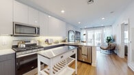This single-floor residence at 1 Midtown Bridge Approach in Hackensack, New Jersey, offers a thoughtfully designed living space characterized by clean lines, modern finishes, and a cohesive contemporary aesthetic. The open-concept kitchen serves as a central highlight, featuring two-tone cabinetry with upper white gloss cabinets and lower charcoal gray panels, complemented by light quartz countertops and a white subway tile backsplash. A full suite of stainless steel appliances—including a French door refrigerator, gas range, microwave, and dishwasher—adds both functionality and visual appeal. Under-cabinet lighting enhances the workspace, while warm hardwood flooring flows throughout, tying the kitchen seamlessly into the living area. Large windows and a glass-paneled balcony door at the far end of the living space invite generous natural light and outdoor access. The bedroom is a calm, well-proportioned retreat with a sizable window, soft blue-gray walls, and hardwood flooring, accompanied by a spacious walk-in closet with built-in shelving and hanging systems. The bathroom presents a clean, spa-like aesthetic with white subway tile surrounding the soaking tub, large-format floor tiles, a wood-toned vanity, and a backlit mirror. Recessed lighting throughout maintains a bright and welcoming ambiance.