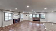 This single-floor space offers an open and versatile layout finished with rich, warm hardwood flooring that flows seamlessly throughout. The main area is generously proportioned, providing ample room for a variety of uses, from social gatherings to event hosting. A dark-framed fireplace with a tile surround anchors one wall, adding a welcoming focal point to the space. Recessed ceiling lighting illuminates the interior evenly, complemented by natural light streaming in through multiple windows fitted with horizontal blinds. The entrance features a striking set of double doors crafted in rich wood tones with decorative leaded glass inserts, creating an elegant and welcoming arrival experience. Wall sconces add subtle ambient lighting alongside framed artwork that enhances the refined atmosphere. Toward the rear, a kitchenette area includes a large island with a dark wood base and light countertop, pendant lighting overhead, a full-size refrigerator, and additional counter space — making the space well-equipped for catering or food service needs. A separate door provides additional access or storage utility. The overall ambiance is clean, neutral, and polished, offering a flexible and attractive environment suitable for a wide range of purposes.