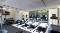 This fitness center offers a clean, contemporary design with a strong emphasis on natural light and an open, airy atmosphere. Floor-to-ceiling glass windows span the length of the space, flooding the interior with sunlight and providing unobstructed views of lush greenery outside — creating an energizing environment for workouts. The white ceiling and neutral-toned walls contribute to the bright, minimalist aesthetic throughout.

The cardio area is well-appointed, featuring a row of modern treadmills aligned along the windowed wall, each equipped with individual digital display screens. Elliptical machines are also positioned nearby, offering a variety of low-impact cardiovascular training options. Additional exercise equipment is visible toward the rear of the space, accommodating a range of fitness needs. The flooring is finished with a dark, resilient material suited for gym use, providing both functionality and contrast against the light interior. Recessed and track lighting supplements the natural light, ensuring the space remains well-lit throughout the day. Overall, the fitness center delivers a polished, professional environment designed for comfort and performance.