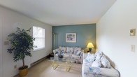 This single-floor residence offers a thoughtfully arranged 1-bedroom, 1-bathroom layout designed for comfortable everyday living. The living room serves as the central gathering space, highlighted by a calming teal accent wall that adds a pop of color against the neutral white walls and ceiling. Plush, floral-patterned seating provides a classic, inviting atmosphere, complemented by a glass-top coffee table with a brass frame that adds a touch of elegance. A floor lamp and table lamp provide warm ambient lighting throughout the room. Natural light enters through a sizable window fitted with horizontal blinds, offering both brightness and privacy control. Baseboard heating runs along the lower walls, ensuring year-round comfort. Carpeted flooring adds warmth and softness underfoot throughout the living area. Decorative touches, including framed wall art and indoor greenery, contribute to the overall homey ambiance. The single-floor design ensures easy accessibility and a practical flow between spaces, making this residence a comfortable and well-appointed living option.