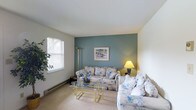 This single-floor residence offers a thoughtfully arranged 1-bedroom, 1-bathroom layout designed for comfortable everyday living. The living room serves as the central gathering space, highlighted by a calming teal accent wall that adds a pop of color against the neutral white walls and ceiling. Plush, floral-patterned seating provides a classic, inviting atmosphere, complemented by a glass-top coffee table with a brass frame that adds a touch of elegance. A floor lamp and table lamp provide warm ambient lighting throughout the room. Natural light enters through a sizable window fitted with horizontal blinds, offering both brightness and privacy control. Baseboard heating runs along the lower walls, ensuring year-round comfort. Carpeted flooring adds warmth and softness underfoot throughout the living area. Decorative touches, including framed wall art and indoor greenery, contribute to the overall homey ambiance. The single-floor design ensures easy accessibility and a practical flow between spaces, making this residence a comfortable and well-appointed living option.