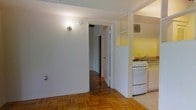 This single-floor residential space offers a practical and classic layout designed for comfortable everyday living. The flooring is a standout feature throughout, with warm parquet hardwood panels adding character and a timeless aesthetic to the main living areas. The space flows naturally between rooms, with a central doorway connecting the main area to an inner hallway, which leads further into the unit and reveals additional rooms beyond.

The kitchen is positioned along one side of the space and is efficiently laid out, featuring a white gas range stove, a sink, and light-colored countertops. Open shelving above provides accessible storage, and the warm yellow-toned walls give the kitchen a bright, welcoming atmosphere. A ventilation unit is mounted on the wall, adding to the kitchen's functional design.

The walls throughout are painted in clean, neutral white tones, contributing to a bright and airy feel. Wall-mounted lighting fixtures provide warm illumination. The hallway glimpsed through the doorway hints at further depth within the space, suggesting a connected series of rooms. Overall, the space presents a no-frills, functional interior with classic residential details that offer a solid foundation for personalization.
