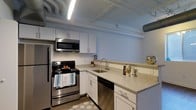 This single-floor space showcases a thoughtfully designed kitchen that serves as its standout feature. The kitchen is outfitted with a full suite of stainless steel appliances, including a refrigerator, electric range with overhead microwave, and dishwasher, providing a complete and functional cooking environment. White shaker-style cabinetry lines the walls, offering ample storage, while the light-toned granite-style countertops add a clean, contemporary aesthetic. The undermount stainless steel sink is positioned at the counter's L-shaped corner, maximizing workspace efficiency on both sides. The flooring features wood-look planks, contributing warmth to the overall design. A distinctive industrial character is introduced through the exposed ceiling, where white-painted pipes, ductwork, and fluorescent tube lighting are left visible overhead — a design choice that adds an urban, loft-inspired aesthetic to the space. The combination of crisp white finishes, modern hardware, and industrial ceiling details creates a balanced contrast between contemporary style and raw architectural character, resulting in a kitchen that is both highly functional and visually engaging.