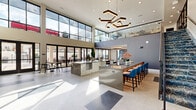 Located at 127 W Moreno Ave in Colorado Springs, CO, this sophisticated two-story amenity space offers a sleek, modern design across a generously sized floor plan. The expansive main level is anchored by a dramatic double-height great room flooded with natural light through floor-to-ceiling glass walls, creating a bright and airy atmosphere. Two fully equipped kitchens serve the space — one features stainless steel islands with bar seating and bold geometric pendant lighting, while the second offers a streamlined wet bar setup with hexagonal tile accents, floating shelves, and integrated beverage refrigerators. Three distinct living areas provide flexible gathering zones, including a lounge with designer patterned carpeting, tufted seating, and an architecturally detailed ceiling with geometric cove lighting. A glass-railed staircase connects to the upper level, which encompasses additional lounge space and provides elevated views of the interior. The balcony, accessible from the main level, features glass railings and overlooks a vibrant courtyard complete with a pool and outdoor seating. Large glass entry doors with dark frames add a refined, contemporary touch throughout. The overall ambiance is polished and inviting, blending modern aesthetics with functional communal design.
