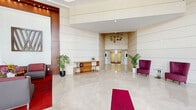 Located at 410 Evernia Street in the heart of West Palm Beach, Florida, this sophisticated lobby space offers a grand and welcoming first impression. The single-floor layout spans a generous footprint, designed to accommodate both movement and comfort with ease.

Upon entry, a dramatic revolving glass door opens into a sun-drenched reception area framed by soaring double-height ceilings and floor-to-ceiling windows that flood the space with natural light and offer views of the surrounding urban streetscape. The highly polished white marble flooring reflects light throughout, enhancing the sense of openness and luxury.

The interior is divided into distinct functional zones. A formal seating area features a glass-top dining table surrounded by rich leather chairs, positioned beneath a wall of tall, multi-pane windows that frame outdoor palm tree views. Adjacent, a lounge area is furnished with contemporary gray upholstered chairs arranged around a red area rug, anchored by a bold geometric wall-mounted art piece set within warm wood wall paneling with recessed accent lighting.

Deeper into the lobby, a pair of gold-finished elevator doors serves as a focal point, flanked by lush tropical potted plants and accented by a striking colorful glass orb wall installation overhead. Purple wingback accent chairs and a sleek open console table add refined pops of color. The overall ambiance is polished, modern, and inviting.