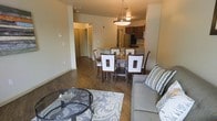 This single-floor residence showcases an open-concept design that seamlessly connects the living, dining, and kitchen areas into one cohesive and functional space. Dark hardwood-style flooring runs throughout, providing a warm contrast against the neutral beige walls and white trim.

The living area is anchored by a plush gray sofa and a round glass-top coffee table positioned over a patterned area rug, creating an inviting and comfortable gathering space. A framed artwork piece adorns the wall, adding a touch of color to the understated palette.

Adjacent to the living area, the dining space features a dark wood table surrounded by upholstered chairs with white accent panels, illuminated by a pendant light fixture that adds a contemporary focal point overhead.

The kitchen is visible beyond the dining area, equipped with dark wood cabinetry, stainless steel appliances including a refrigerator, and a functional layout designed for everyday use. A hallway leading to additional rooms is visible in the background, suggesting a practical and well-organized floor plan. Overall, the space presents a modern, clean aesthetic with cohesive finishes throughout.