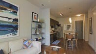 This single-floor residence offers a thoughtfully designed open-concept layout that integrates the living, kitchen, and dining areas into one cohesive and functional space. The living area is anchored by a clean, minimalist aesthetic, featuring rich hardwood-style flooring that extends throughout and lends warmth to the interior. A stylish metal open-shelf bookcase serves as a subtle room divider, adding a decorative yet practical element to the space.

The kitchen is well-appointed with dark cabinetry, stainless steel appliances including a refrigerator, range, and dishwasher, and is illuminated by modern track lighting overhead. A mosaic-tile runner adds a charming accent on the kitchen floor. Adjacent to the kitchen, the dining area is defined by a pendant light fixture and comfortably accommodates a dining table with modern wooden chairs, making it ideal for both casual meals and entertaining.

The space benefits from a neutral color palette across the walls and ceilings, creating a bright and airy ambiance. Visible ceiling vents suggest a central HVAC system for year-round comfort. Overall, the unit presents a contemporary, urban lifestyle aesthetic that is both stylish and highly livable.
