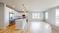 This 870-square-foot, one-bedroom, one-bathroom residence at 1211 Carrington Blvd in Mount Pleasant, WI offers a clean, modern aesthetic with high-quality finishes throughout its single-floor layout. The open-concept kitchen and living area serves as the heart of the unit, featuring a large central island with a white quartz countertop, dual-tone cabinetry with white upper and gray lower shaker-style cabinets, matte black hardware, and stainless steel appliances including a range, microwave, and French door refrigerator. Pendant lighting and light hardwood flooring complete the stylish kitchen. The living room is bright and airy, with multiple windows and a glass sliding door leading to a private outdoor balcony. The 150-square-foot bedroom is carpeted and well-lit by large windows offering neighborhood views, with two doors providing access to the hallway and closet. The bathroom features a gray vanity with white countertop, matte black fixtures, a large mirror, and hardwood-style flooring. Throughout the unit, neutral warm-gray walls, light hardwood flooring, and an abundance of natural light create a cohesive, contemporary atmosphere.