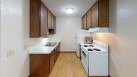 Located at 825 9th Ave S in Saint Cloud, Minnesota, this single-floor unit offers a practical and functional layout spread across several well-defined living spaces. The open entry area transitions seamlessly between the kitchen and the main living space, with light hardwood-style flooring anchoring the more active areas of the unit. The galley-style kitchen is equipped with rich dark wood cabinetry offering ample storage above and below the countertops, white appliances including a coil-top range and refrigerator, and a double-basin sink set into clean white countertops — all beneath a textured ceiling with flush lighting. The living area is generously proportioned, featuring neutral carpet, baseboard heating, a wall-mounted air conditioning unit, and a sliding glass door dressed with vertical blinds that invites natural light and outdoor access. Two bedrooms mirror each other in design, each offering carpeted floors, a window with vertical blinds providing natural light, baseboard heating, and textured ceilings. The full bathroom includes a single-sink vanity with dark wood cabinetry, a toilet, and a shower-tub combination with curtain, all set atop classic mosaic tile flooring. The unit's neutral color palette and consistent finishes create a cohesive, move-in-ready environment.