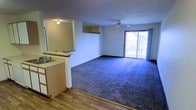 This single-floor residential space is designed with an open-concept layout that seamlessly connects the kitchen and living areas. The kitchen features wood-toned cabinetry, a built-in sink, and a pass-through window that opens directly into the main living space, making it ideal for entertaining or everyday convenience. A transition from wood-style hard flooring in the kitchen leads into the plush carpeted living area, providing a comfortable and cozy atmosphere throughout.

The spacious living room is highlighted by a ceiling fan that promotes year-round air circulation, and a textured ceiling that adds subtle architectural character. A large sliding glass door dominates one wall of the living area, allowing abundant natural light to fill the room while offering a view of the surrounding exterior. Curtain panels frame the door, providing flexibility for privacy and light control.

The neutral wall tones and understated finishes throughout create a clean, functional aesthetic that offers a versatile backdrop for a variety of interior styles. The single-floor layout ensures easy accessibility and a cohesive flow between all areas of the space.