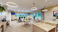 Located at 1000 Cowpen Ct in Williamsburg, Virginia, this single-floor communal space offers a thoughtfully designed open-concept layout that seamlessly blends a fully equipped kitchen with a spacious living and lounge area. The entrance greets visitors with a striking marble-patterned tile floor accented by wood inlays, elegant wall décor, and linear LED cove lighting that runs along the ceiling to create a polished, contemporary atmosphere. Structural columns and clean architectural lines define the transition between zones while maintaining an open, airy feel throughout.

The kitchen area is appointed with expansive granite countertops, light wood cabinetry, stainless steel appliances including a full-size refrigerator, an under-counter unit, and a sink — all complemented by wall-mounted flat-screen TVs and modern pendant lighting. Stylish blue stools line the island, making it ideal for casual seating and social interaction.

The living area transitions to plush carpeted flooring and is furnished with contemporary sofas, lounge chairs, and multiple dining tables with chairs arranged to encourage gathering. An array of large windows along the perimeter floods the space with natural light and offers lush green outdoor views. Decorative sculptural elements, globe-shaped topiaries, and curated artwork enhance the welcoming, refined ambiance throughout.