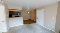 This single-floor residential unit offers a functional and welcoming open-concept layout that seamlessly connects the main living areas. The living room is finished with soft, neutral-toned carpeting that adds warmth underfoot, while the adjoining dining area transitions to smooth hardwood-style flooring, creating a natural distinction between the two spaces. A pendant light fixture hangs above the dining area, providing a focal point and practical illumination for the space.

The kitchen is positioned adjacent to the living and dining areas, separated by a raised breakfast bar counter finished with a granite-style surface, offering both a casual dining option and a natural visual divider. Dark espresso-toned cabinetry lines the kitchen walls, complemented by black appliances including a built-in microwave and range, lending a cohesive and modern look to the cooking space.

Throughout the unit, warm beige walls create a neutral and inviting backdrop that pairs well with both the cabinetry and flooring choices. White trim and baseboards provide clean contrast along the walls and doorways. A classic paneled white entry door adds a traditional architectural detail to the space. The overall ambiance is clean, practical, and move-in ready, well-suited for comfortable everyday living.
