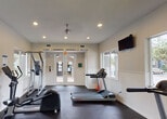 This fitness center at 4495 Emerald Vista in West Palm Beach, Florida, is a well-equipped, single-story facility designed to support a comprehensive workout experience. The space is organized into distinct cardio and strength training zones, each thoughtfully arranged to maximize usability and flow.

The cardio area is outfitted with multiple treadmills, elliptical machines, a stationary bike, and a stair climber, providing a wide range of aerobic training options. A wall-mounted flat-screen television is positioned for convenient viewing during workouts. Recessed lighting throughout the space ensures consistent illumination, while several windows and glass double doors allow natural light to fill the room and offer views of the surrounding outdoor landscape, including glimpses of a pool area and green grounds.

The strength training section features a variety of cable and selectorized resistance machines targeting multiple muscle groups, accommodating users of varying fitness levels. The durable rubber flooring throughout the facility provides traction and impact absorption across both zones.

The interior maintains a clean, neutral aesthetic with white walls and a smooth ceiling. Multiple exit points with glass-panel double doors connect the space directly to the outdoors, enhancing ventilation and accessibility. Overall, the fitness center presents a functional and well-maintained environment suited for daily exercise.