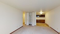 This single-floor space offers a clean, practical layout designed to maximize functionality. The main living area features neutral wall-to-wall carpeting that lends a quiet, understated warmth to the room. A prominent corner closet with bi-fold doors provides generous built-in storage, complete with interior shelving and a hanging rod — ideal for organization. Adjacent to the closet, a hallway leads further into the space, suggesting additional rooms or utility areas beyond the immediate view. Transitioning from the carpeted area, the flooring shifts to a classic tile pattern, delineating a secondary zone that features rich, dark wood cabinetry along the lower wall and open shelving above. A flush-mounted ceiling light illuminates this area, highlighting the warm tones of the woodwork. The white walls and light ceiling throughout contribute to a bright, airy atmosphere, while the baseboard heating along the perimeter reflects the space's practical, well-considered design. Overall, the space presents a straightforward, functional character with thoughtful built-in features that offer ample storage and a clear, organized flow between zones.