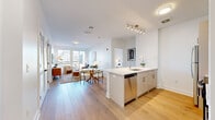 This thoughtfully designed single-floor residence offers a bright, contemporary living experience defined by clean lines, neutral tones, and quality finishes. The open-concept layout seamlessly connects the kitchen, dining, and living areas, all unified by wide-plank light hardwood flooring that extends throughout the main living spaces. The kitchen is a standout feature, anchored by a large waterfall-edge quartz island with bar seating and an integrated sink. Crisp white shaker cabinetry, a geometric tile backsplash, and a full suite of stainless steel appliances complete the well-equipped space. The living room is bathed in natural light from oversized windows that frame urban views, creating an airy, inviting atmosphere. Both bedrooms are generously proportioned, each featuring large windows with city views and light hardwood floors. The bathrooms are finished to a high standard, with the primary bath showcasing a quartz-topped vanity with brushed gold fixtures and a glass-enclosed walk-in shower clad in large-format neutral tile with brushed nickel hardware. The overall aesthetic throughout the residence is modern and cohesive, with white walls, recessed and track lighting, and refined material choices creating a polished yet welcoming environment.