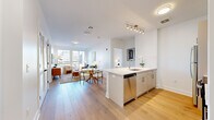 This thoughtfully designed single-floor residence offers a bright, contemporary living experience defined by clean lines, neutral tones, and quality finishes. The open-concept layout seamlessly connects the kitchen, dining, and living areas, all unified by wide-plank light hardwood flooring that extends throughout the main living spaces. The kitchen is a standout feature, anchored by a large waterfall-edge quartz island with bar seating and an integrated sink. Crisp white shaker cabinetry, a geometric tile backsplash, and a full suite of stainless steel appliances complete the well-equipped space. The living room is bathed in natural light from oversized windows that frame urban views, creating an airy, inviting atmosphere. Both bedrooms are generously proportioned, each featuring large windows with city views and light hardwood floors. The bathrooms are finished to a high standard, with the primary bath showcasing a quartz-topped vanity with brushed gold fixtures and a glass-enclosed walk-in shower clad in large-format neutral tile with brushed nickel hardware. The overall aesthetic throughout the residence is modern and cohesive, with white walls, recessed and track lighting, and refined material choices creating a polished yet welcoming environment.