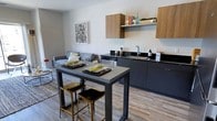 This single-floor residence showcases a thoughtfully designed open-concept layout where the living area and kitchen flow seamlessly together. The living space features large sliding glass doors that invite abundant natural light and provide views of the surrounding urban streetscape, complemented by hardwood-style flooring and a textured area rug anchoring the seating arrangement.

The kitchen is outfitted with dark charcoal cabinetry paired with warm wood-toned upper cabinets, black granite countertops, and stainless steel appliances including a range, microwave, dishwasher, and refrigerator. A high-top dining table with bar-height seating bridges the kitchen and living areas, making the space ideal for both casual dining and entertaining.

The unit includes two bedrooms. The primary bedroom features multiple windows with sheer curtains that fill the room with soft, diffused light. The second bedroom maintains the same bright, airy quality with a clean, minimalist aesthetic. Both rooms offer comfortable accommodations with ample natural light and urban views.

The bathroom presents a cohesive design with a dark-framed vanity mirror, sleek white sink set into dark cabinetry, and a glass-enclosed walk-in shower with built-in shelving niches, all finished in a cool blue-grey palette.