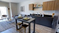 This single-floor residence showcases a thoughtfully designed open-concept layout where the living area and kitchen flow seamlessly together. The living space features large sliding glass doors that invite abundant natural light and provide views of the surrounding urban streetscape, complemented by hardwood-style flooring and a textured area rug anchoring the seating arrangement.

The kitchen is outfitted with dark charcoal cabinetry paired with warm wood-toned upper cabinets, black granite countertops, and stainless steel appliances including a range, microwave, dishwasher, and refrigerator. A high-top dining table with bar-height seating bridges the kitchen and living areas, making the space ideal for both casual dining and entertaining.

The unit includes two bedrooms. The primary bedroom features multiple windows with sheer curtains that fill the room with soft, diffused light. The second bedroom maintains the same bright, airy quality with a clean, minimalist aesthetic. Both rooms offer comfortable accommodations with ample natural light and urban views.

The bathroom presents a cohesive design with a dark-framed vanity mirror, sleek white sink set into dark cabinetry, and a glass-enclosed walk-in shower with built-in shelving niches, all finished in a cool blue-grey palette.