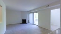 This single-floor living space offers a clean, understated aesthetic defined by neutral tones and practical design. The main living area is carpeted in a soft, light gray that extends throughout, creating a cohesive and quiet atmosphere. White walls and a white ceiling contribute to the overall brightness of the space, making it feel open and airy.

A fireplace with a dark surround serves as a subtle focal point along one wall, providing both visual interest and functional warmth. Adjacent to the fireplace, a wall-mounted heating unit ensures efficient climate control.

Natural light plays a prominent role in the space, courtesy of a full-height sliding glass door fitted with vertical blinds. This door opens to an outdoor balcony, where a railing is visible beyond the glass, suggesting an elevated position with an outdoor view of surrounding greenery. The sliding door allows for an easy transition between the interior and exterior while flooding the room with daylight.

The overall design is minimal and move-in ready, with architectural details kept simple and clean. The single-floor layout offers straightforward, accessible living with a calm, functional ambiance throughout.