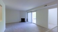 This single-floor living space offers a clean, understated aesthetic defined by neutral tones and practical design. The main living area is carpeted in a soft, light gray that extends throughout, creating a cohesive and quiet atmosphere. White walls and a white ceiling contribute to the overall brightness of the space, making it feel open and airy.

A fireplace with a dark surround serves as a subtle focal point along one wall, providing both visual interest and functional warmth. Adjacent to the fireplace, a wall-mounted heating unit ensures efficient climate control.

Natural light plays a prominent role in the space, courtesy of a full-height sliding glass door fitted with vertical blinds. This door opens to an outdoor balcony, where a railing is visible beyond the glass, suggesting an elevated position with an outdoor view of surrounding greenery. The sliding door allows for an easy transition between the interior and exterior while flooding the room with daylight.

The overall design is minimal and move-in ready, with architectural details kept simple and clean. The single-floor layout offers straightforward, accessible living with a calm, functional ambiance throughout.
