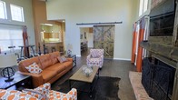 This single-story space showcases a thoughtfully designed open floor plan that emphasizes both functionality and character. The living area is the centerpiece of the space, boasting impressively high ceilings that amplify the sense of openness and airiness. Dark hardwood-style flooring runs throughout, providing a rich, grounding contrast to the light-toned walls.

A standout architectural feature is the large, weathered wood sliding barn door mounted on an industrial black rail, adding a rustic-chic aesthetic that anchors the space with visual interest. The living room is furnished with a warm cognac leather sofa and coordinating patterned accent chairs in orange and white tones, creating a cohesive and inviting seating arrangement around a wood-and-metal coffee table.

Through an open archway, the kitchen is visible and features bar-height seating along a counter, pendant lighting, and warm cabinetry, seamlessly integrating dining and entertaining areas. A glimpse of a formal dining space with additional seating is visible beyond the barn door. Natural light filters into the space, enhancing the warm, welcoming ambiance throughout.