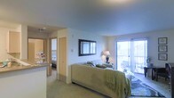 This single-floor residential space is thoughtfully designed with an open, flowing layout that connects multiple living areas. The main bedroom area is a comfortable, well-proportioned space featuring plush carpeted flooring and soft, neutral-toned walls that create a calm and welcoming atmosphere. A framed decorative mirror mounted on the wall adds a classic touch, while a stylish table lamp provides warm ambient lighting. A sliding glass door leads directly to a private balcony, allowing natural light to fill the room and offering an outdoor retreat. The balcony appears to feature seating, suggesting a pleasant area for relaxation. Adjacent to the main bedroom area, a closet with a light wood-toned door provides practical storage. A hallway visible in the background connects to an additional bedroom, also carpeted, hinting at a multi-room configuration. To one side, a kitchen counter or bar area is partially visible, indicating an integrated living and kitchen space. Overall, the space presents a balanced combination of functionality and comfort, with consistent neutral finishes, soft lighting, and a cohesive design aesthetic throughout.