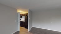 This single-floor residential unit offers a practical and well-organized layout that makes efficient use of its space. The living area is finished with durable wood-look vinyl flooring in a warm gray tone, neutral gray walls, and clean white baseboards, creating a bright and cohesive atmosphere. A large window fitted with vertical blinds allows natural light to fill the room while offering outdoor views of surrounding greenery. A baseboard heater runs along the exterior wall, providing efficient climate control.

The kitchen is positioned just off the living area and is accessible through an open doorway. It features rich dark espresso cabinetry — both upper and lower — providing ample storage. The kitchen is equipped with black appliances including a coil-burner electric range and a full-size refrigerator, complemented by a single-basin stainless steel sink set into a light-toned countertop.

The bedroom offers a quiet, well-proportioned space with the same dark wood-look flooring and neutral wall tones, providing a calm and private retreat.

The bathroom includes a full-size soaking tub with a tub/shower combination, finished with crisp white surround panels and a built-in niche for storage. Chrome fixtures complete the clean, functional aesthetic throughout the space.