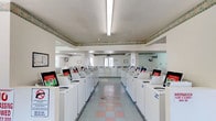 This single-floor laundromat offers a clean, well-organized environment designed to accommodate a high volume of laundry needs with ease. The facility is lined with rows of commercial-grade washing machines and dryers arranged symmetrically on both sides of a central aisle, providing ample capacity for simultaneous use. The machines are uniformly white and clearly labeled, contributing to the tidy, orderly appearance of the space.

The interior is finished with light-colored walls accented by a decorative border running along the upper perimeter, adding a subtle touch of character to the otherwise utilitarian setting. The flooring features a classic checkered tile pattern in muted pastel tones, lending a retro charm to the space. Overhead, bright fluorescent lighting ensures the entire facility is well-lit throughout the day.

Natural light filters in through windows positioned at the far end of the room, enhancing the bright and airy atmosphere. Clocks are mounted on the walls for the convenience of patrons keeping track of their laundry cycles. The layout is thoughtfully designed with a wide central walkway, allowing for comfortable movement between machines. Overall, this laundromat presents a practical, clean, and user-friendly environment suited for everyday laundry needs.