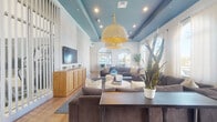 Located at 11700 SW 26th St in Miramar, Florida, this single-floor amenity space is thoughtfully designed to offer residents a refined and functional communal environment. The centerpiece of the space is a spacious lounge area distinguished by its striking teal-painted tray ceiling, warm wood flooring, and a statement woven pendant light. Generous arched windows draped in sheer white curtains flood the room with natural light while framing views of the surrounding community, creating an airy and welcoming atmosphere. Comfortable sectional seating arrangements and a wall-mounted flat-screen TV make this lounge ideal for both relaxation and socializing. Adjacent to the lounge, a dedicated meeting and dining area features wood-look flooring, a white dining table with mixed seating, a wall-mounted display screen, and tall ceilings that enhance the sense of openness. The wide corridors connecting the spaces are finished with large-format travertine-style tile and accented by sage green walls and curated artwork, lending a polished, gallery-like quality. Floor-to-ceiling glass walls reveal a well-equipped fitness center alongside the corridor, offering visibility into the workout space. A separate business or co-working nook with pendant lighting rounds out the amenities, making this a comprehensive and stylish community facility.