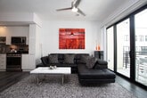 This contemporary single-floor residence showcases a thoughtfully designed open-concept layout with clean lines and a modern aesthetic throughout. The living area is defined by wood-look flooring, a ceiling fan, and floor-to-ceiling sliding glass doors that open to a private balcony with outdoor views. A striking frosted glass and black-framed sliding partition elegantly separates the living space from the bedroom, allowing natural light to pass through while providing privacy when needed.

The bedroom accommodates a full-size bed and includes a ceiling fan, a window with exterior views, and direct visual connection to the balcony through the glass partition. The galley-style kitchen is well-appointed with high-gloss white cabinetry extending to the ceiling, stainless steel appliances including a gas range, built-in microwave, dishwasher, and refrigerator, complemented by quartz countertops and a neutral tile backsplash.

The bathroom features large-format grey tile flooring, a floating vanity with an undermount sink and grey countertop, a wall-mounted toilet, and a glass-enclosed walk-in shower with a rain showerhead. The overall ambiance is clean, minimal, and contemporary.