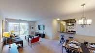 This single-story residence offers a thoughtfully designed, open-concept layout that connects the main living spaces seamlessly. The carpeted living room is generously sized, featuring a wall-mounted flat-screen TV, a sliding glass door with vertical blinds, and direct access to an outdoor patio with exterior views. Adjacent to the living room, a dedicated dining area features a pendant chandelier overhead and flows naturally into the kitchen via a breakfast bar with bar stool seating.

The kitchen is well-equipped with warm wood cabinetry, dark granite-style countertops, and a full suite of black appliances including a refrigerator, electric range, built-in microwave, and dishwasher. Light wood-style flooring ties the kitchen together and contrasts with the carpet found throughout the rest of the unit.

The bedroom is notably spacious, featuring plush carpet, large windows with sheer curtains that allow natural light, and two closets offering generous storage. The full bathroom includes a tub-shower combination with tile surround and wood-style flooring, accessible via a short hallway from the bedroom. Overall, the space presents a clean, neutral palette with a functional and comfortable layout.