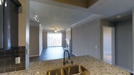 This single-story residence in Plano, Texas, showcases an open-concept design that seamlessly connects the kitchen and living areas. The kitchen features striking granite countertops with warm gold and beige tones, complemented by a double-basin stainless steel sink equipped with a modern pull-down faucet. Track lighting overhead adds focused illumination to the kitchen workspace.

The layout opens directly into a spacious living area, which is enhanced by crown molding that adds a refined architectural detail along the ceiling perimeter. A classic white fireplace serves as a focal point in the living room, providing both a functional and decorative element. Large windows dressed with vertical blinds allow generous natural light to fill the space, brightening the room throughout the day.

Dark hardwood-style flooring runs through the living area, creating a warm contrast against the neutral gray walls. A ceiling fan with a light fixture provides both comfort and supplementary lighting. The overall ambiance is clean and contemporary, with a neutral color palette and thoughtful finishes that create a welcoming, well-appointed living environment.
