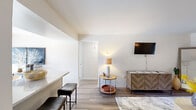 This single-floor, 3-bedroom, 1-bathroom residence offers a thoughtfully arranged layout that makes efficient use of its space. The living room features hardwood-style flooring, neutral white walls, and a wall-mounted television above a chevron-patterned media console, creating a clean and modern atmosphere. Natural light filters in through a window dressed with curtains, adding warmth to the room.

The combined dining and kitchen area is a standout feature of the unit. The dining space is illuminated by a ceiling fan with light fixture and benefits from an abundance of natural light through full-length sliding glass doors fitted with vertical blinds, offering views of the outdoors. The kitchen is equipped with gray cabinetry, light countertops, and full-sized appliances, and flows openly into the adjacent living and dining areas.

The primary bedroom is a comfortable retreat, featuring carpeted flooring, a large window with vertical blinds and drape panels, and ample wall space. The shared bathroom is finished with white tile, a white vanity with chrome fixtures, a large mirror with Hollywood-style lighting, and a shower-tub combination. Additional storage is provided by a dedicated closet with a full hanging rod and shelf. A hallway connects the bedrooms and serves as a functional transition space throughout the unit.