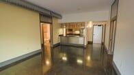 This single-floor unit showcases a striking industrial aesthetic defined by gleaming polished concrete floors that reflect ambient light throughout the open living area. The layout flows seamlessly from the main living space toward a centrally positioned kitchen, which serves as a functional focal point. The kitchen features warm wood cabinetry with glass-front upper cabinets, stainless steel appliances including a microwave and refrigerator, a tile backsplash, and a distinctive peninsula with eye-catching diamond-plate metal paneling and a dark countertop — a nod to the space's industrial character. Exposed ductwork runs along the ceiling, reinforcing the loft-inspired design language. The unit's neutral-toned walls and recessed lighting keep the interior bright and versatile. A sliding industrial-style door on the left side of the space provides access to a secondary area, which includes a visible bathroom with a toilet and refrigerator. A private entry door is positioned to the right, completing the layout. The overall ambiance balances raw, urban materials with functional design, creating a distinctive and contemporary living environment.