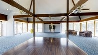This single-story space makes a striking impression with its dramatic vaulted ceiling supported by bold, dark-stained exposed wood beam trusses that form a distinctive architectural framework overhead. The generous ceiling height creates an open, airy atmosphere that adds to the overall sense of spaciousness. The interior is defined by a transition between rich, dark hardwood flooring in the foreground and plush carpeting in the rear portion of the space, subtly delineating functional zones. Panoramic windows line multiple walls, flooding the interior with natural light and offering views of the surrounding outdoor landscape. A ceiling fan is centrally mounted to aid in climate comfort beneath the soaring roofline. The rear area features a pair of potted trees flanking a central focal point, adding a natural, decorative element to the space. Wood paneling along the mid-wall sections complements the exposed timber above, reinforcing the warm, rustic character of the design. The overall ambiance is open and welcoming, with a blend of natural materials and abundant light creating a versatile and inviting environment suitable for a variety of uses.
