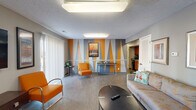 Located at 201 W Saint Clair St in Vincennes, Indiana, this single-floor community space combines bold design with functional versatility. The main lounge area makes an immediate impression with its striking accent wall featuring large geometric triangles in vivid orange and gray — a defining visual centerpiece of the space. The lounge is furnished with a full sofa, retro-style orange accent chairs, and a round coffee table, all set atop durable wood-look flooring. A flat-screen TV is mounted on the wall, and sliding glass doors allow natural light to filter in. Adjacent to the lounge, a compact kitchen is visible through an open passageway, equipped with warm wood cabinetry, a black refrigerator, and a sink with countertop workspace. A separate waiting or reception nook features swivel lounge chairs in neutral gray against a warm terracotta accent wall, alongside a small workstation area. The casual dining section offers high-top bistro tables and bar stools arranged near large windows that flood the room with natural light. A popcorn machine and additional seating add a recreational, welcoming touch to this area. Throughout, the textured ceilings, framed artwork, and cohesive color palette of orange, gray, and taupe create a lively yet approachable atmosphere suited for community gatherings and everyday use.