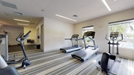 This fitness center is organized across a single floor and divided into two distinct workout zones, offering a comprehensive exercise environment. The cardio area features multiple treadmills and a stationary bike positioned on striped, cushioned carpeting, complemented by large windows that allow abundant natural light and views of the surrounding lush, tropical landscaping. Linear LED lighting runs along the ceiling, keeping the space bright and energizing. The strength training area is outfitted with rubber flooring for durability and safety, and includes a cable machine, a lat pulldown station, a rowing machine, a weight bench, and stability balls for functional training. A wall-mounted television is positioned for viewing during workouts. Ceiling fans in the strength zone provide additional air circulation. The space also features glass doors that open to an outdoor walkway, further enhancing ventilation and connectivity to the exterior grounds. Additional doors along the back wall provide access to adjacent rooms or amenities. The overall design is clean and functional, with neutral-toned walls and practical finishes throughout, making it a well-rounded facility suited for a variety of fitness routines.