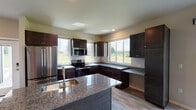 This single-floor home offers a thoughtfully designed open-concept layout that seamlessly connects the kitchen and living area. The kitchen is a standout feature, showcasing rich dark cabinetry, granite countertops, a center island with a built-in sink, and a full suite of stainless steel appliances including a refrigerator, range, and over-the-range microwave. Multiple windows flood the kitchen with natural light and offer views of the surrounding greenery. Light-toned luxury vinyl plank flooring runs throughout the home, providing a clean, cohesive look. A sliding glass door in the living area extends the space visually and provides direct access to the outdoors. The bedrooms are bright and well-sized, each featuring ceiling fans, large windows with open landscape views, and baseboard heating. Closet space is generous, with walk-in closets equipped with wire shelving systems for organized storage. The bathrooms are finished with matching dark cabinetry and granite vanity tops. One bathroom features an accessible roll-in shower with grab bars, while another offers a full vanity and toilet in a practical layout. The overall design balances modern finishes with functional, comfortable living across every room.
