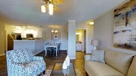 This single-floor unit showcases an open-concept layout that seamlessly connects the living, dining, and kitchen areas into one cohesive space. The living area is anchored by a ceiling fan with a light fixture and features hardwood-style flooring that extends throughout, adding warmth and visual continuity. A glass-top coffee table and a mix of patterned and neutral upholstered seating contribute to a relaxed yet polished atmosphere.

The dining area sits adjacent to the living space, furnished with a classic white dining table and coordinating chairs beneath a decorative chandelier. A wall clock adds a subtle decorative accent to the dining nook. Beyond, the kitchen is partially visible, offering white cabinetry, countertops, and stainless steel appliances, including a microwave.

A structural column subtly defines the transition between the living and dining zones. Toward the rear of the unit, a hallway leads to a bathroom featuring white cabinetry and a well-lit vanity mirror. The textured ceiling and neutral gray walls throughout provide a clean, understated backdrop that ties the entire space together.