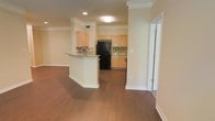 This single-floor residence offers a thoughtfully designed open-concept layout that seamlessly connects the living area to the kitchen. Rich hardwood-style flooring flows throughout the main living space, complemented by crisp white baseboards and elegant crown molding that adds a refined architectural touch to every room.

The kitchen is a standout feature, showcasing light maple cabinetry that provides ample storage, a decorative tile backsplash, and granite or stone-look countertops. A peninsula with a built-in sink extends into the space, creating a natural divide between the kitchen and living area while offering additional prep and seating space. Black appliances, including a full-size refrigerator, anchor the kitchen with a bold contrast against the warm cabinetry.

Structural columns with detailed millwork frame the kitchen entrance, enhancing the sense of definition between spaces without sacrificing the open feel. Recessed and flush-mount ceiling lighting ensures the space is well-lit, while neutral warm tones on the walls create a welcoming and versatile ambiance. A hallway visible from the main area suggests additional rooms beyond, making this a functional and comfortable living environment suited for everyday living.