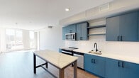 This single-floor unit showcases a thoughtfully designed open layout with consistent hardwood-style flooring connecting the main living areas. The kitchen is a standout feature, outfitted with striking steel-blue cabinetry accented by matte black hardware, white subway tile backsplash, and bright white quartz countertops. A full suite of stainless steel appliances — including a refrigerator, electric range, built-in microwave, and dishwasher — completes the space. A freestanding kitchen island with a matching quartz top and industrial-style base provides additional prep and dining space. Open shelving adds a modern touch alongside the upper cabinetry. The living area is bathed in natural light through expansive floor-to-ceiling windows that offer urban views, creating an airy, open atmosphere. A louvered closet door and additional room access are visible, indicating well-organized storage and a separate bedroom. The bathroom features a clean, minimalist design with speckled tile flooring, a wood-toned vanity with an undermount sink, and accessibility-friendly grab bars. Recessed lighting and neutral wall tones throughout contribute to the space's modern, cohesive aesthetic.