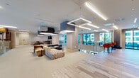 Located at 150 River St in Hackensack, NJ, this single-floor commercial amenity space showcases a thoughtfully designed, open-plan layout that balances collaborative and private zones. The main lounge area features plush, modular seating in warm neutral and earthy tones, anchored by bold geometric pendant lights and acoustic ceiling fixtures suspended above light-toned carpet. Wood-look plank flooring defines transitional corridors and the dining zone, lending warmth and visual contrast throughout.

A café-style lounge area includes booth seating set against a striking hexagonal patterned accent wall, paired with a stone-topped counter bar and pendant lighting in teal and green. Large floor-to-ceiling windows line the perimeter, drawing in generous natural light and providing outdoor views.

Glass-enclosed conference and meeting rooms, framed in sleek black steel, offer transparency while maintaining acoustic separation. Vivid accent furnishings—orange lounge chairs, red stools, and colorful ottomans—punctuate the otherwise neutral palette. Curated artwork adorns the walls throughout.

An interior overlook with a brass-railed glass balustrade reveals a marble-floored lobby atrium below, featuring dramatic pendant lighting and a grand glazed entry, reinforcing the building's polished, contemporary character.