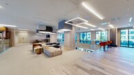 Located at 150 River St in Hackensack, NJ, this single-floor commercial amenity space showcases a thoughtfully designed, open-plan layout that balances collaborative and private zones. The main lounge area features plush, modular seating in warm neutral and earthy tones, anchored by bold geometric pendant lights and acoustic ceiling fixtures suspended above light-toned carpet. Wood-look plank flooring defines transitional corridors and the dining zone, lending warmth and visual contrast throughout.

A café-style lounge area includes booth seating set against a striking hexagonal patterned accent wall, paired with a stone-topped counter bar and pendant lighting in teal and green. Large floor-to-ceiling windows line the perimeter, drawing in generous natural light and providing outdoor views.

Glass-enclosed conference and meeting rooms, framed in sleek black steel, offer transparency while maintaining acoustic separation. Vivid accent furnishings—orange lounge chairs, red stools, and colorful ottomans—punctuate the otherwise neutral palette. Curated artwork adorns the walls throughout.

An interior overlook with a brass-railed glass balustrade reveals a marble-floored lobby atrium below, featuring dramatic pendant lighting and a grand glazed entry, reinforcing the building's polished, contemporary character.
