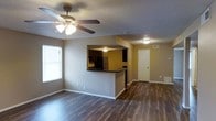 This single-floor space features an open-concept layout that seamlessly connects the living area to the kitchen. The living room is finished with dark hardwood-style flooring that extends throughout, creating a cohesive and grounded aesthetic. A ceiling fan with an integrated light fixture is centrally mounted on the textured ceiling, providing both air circulation and ambient lighting. A large window along one wall allows natural light to fill the space.

The kitchen is positioned adjacent to the living area, separated by a raised breakfast bar with a dark countertop that doubles as a casual dining or prep surface. Visible through the pass-through opening, the kitchen is equipped with dark cabinetry and a stainless steel refrigerator, offering a functional and organized cooking space. Overhead lighting illuminates the kitchen area effectively.

A hallway visible from the living area leads to additional rooms, with a white door providing privacy. The walls are painted in a neutral warm gray tone throughout, complementing the flooring and creating a clean, understated palette. Textured ceilings and white baseboards complete the interior details, lending a practical and tidy finish to the overall space.